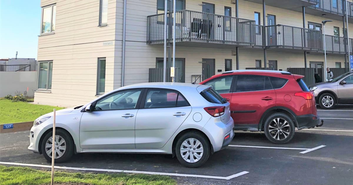 Fyra bilar står på en parkeringsplats framför ett lägenhetshus. Det är en silvrig bil, en röd bil, en guldig bil och en svart bil. Huset är tre våningar, vitt och har balkonger. Himlen är blå.