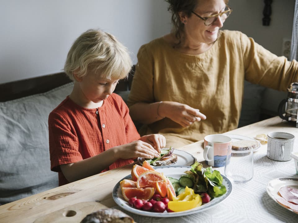 En kvinna och en flicka sitter vid ett träbord med mönstrad vit och grå duk. Det är uppdukat för frukost. På bordet finns ett stort fat med frukt och grönsaker.