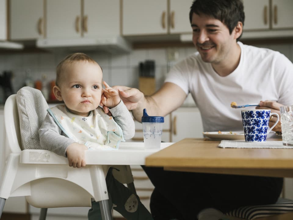 En pappa sitter med sitt småbarn vid ett matbord. Barnet sitter i en  barnstol. Pappa försöker mata barnet, men barnet tittar bort.