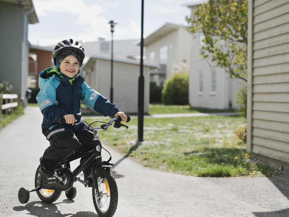En pojke med hjälm på huvudet cyklar runt i ett bostadsområde. Han ser glad ut.