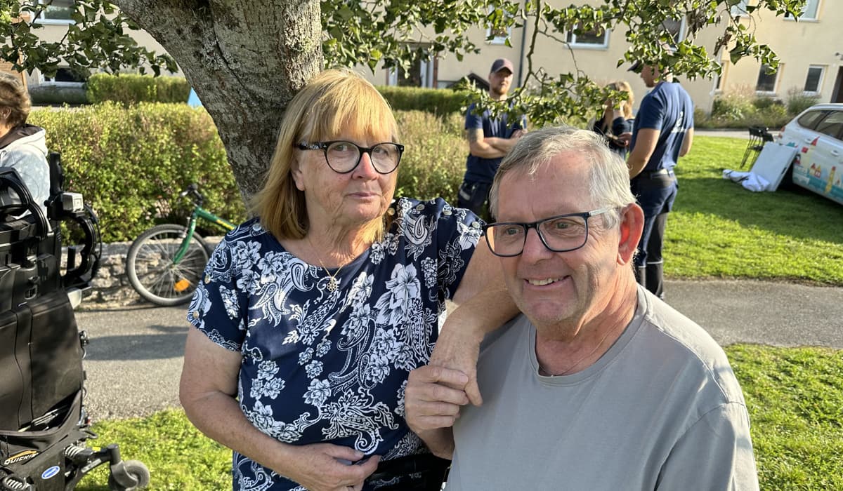 En kvinna med rött hår och svarta glasögon håller en man i handen. Mannen har grå t-shirt och ler