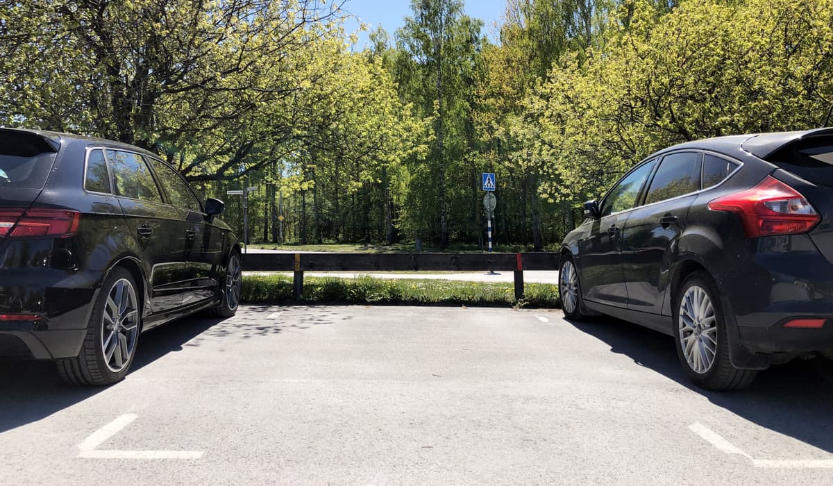 Parkeringsplats med tre parkeringar. Till vänster och höger i bild står bilar parkerade, de är fotade bakifrån. I mitten är det en tom parkeringsplats. Framför bilarna är det träd och grönska.