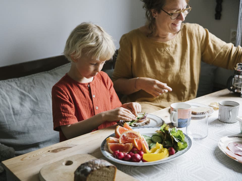 En kvinna och en flicka sitter vid ett träbord med mönstrad vit och grå duk. Det är uppdukat för frukost. På bordet finns ett stort fat med frukt och grönsaker.