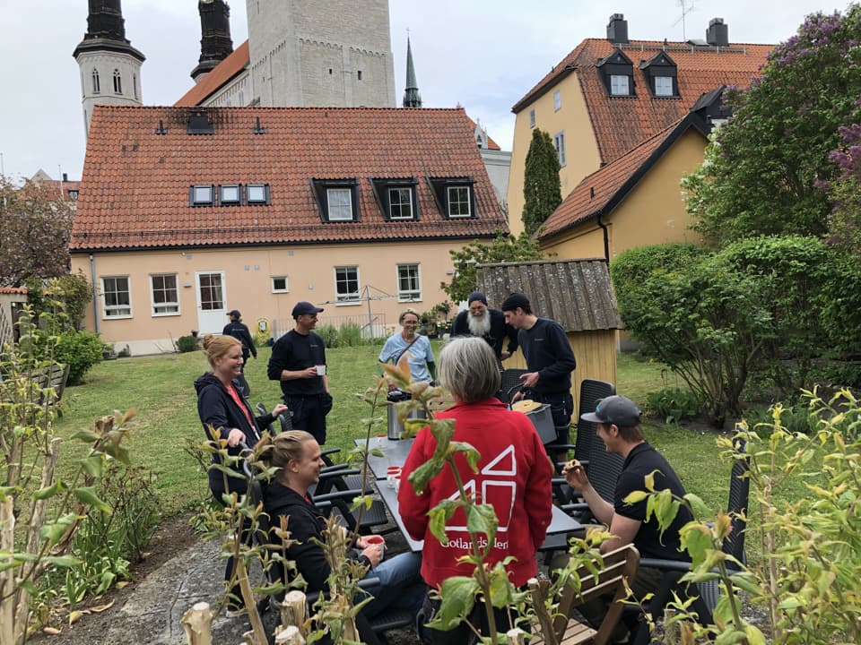 Åtta personer umgås kring ett bord, några sitter och några står. De har kaffekoppar, termos och fika. De sitter på en uteplats i en trädgård bland hus. I bakgrunden syns Visby Domkyrka. 