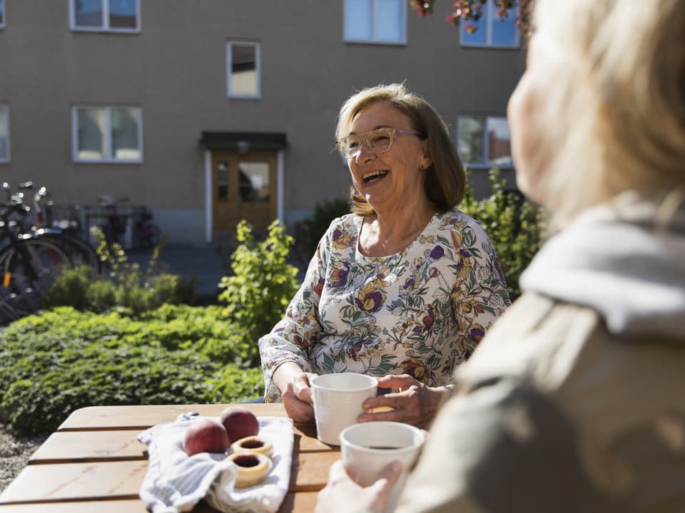 Två kvinnor sitter vid ett bord utomhus och fikar. Den ena kvinnan syns bara bakifrån och den andra kvinnan skrattar. De sitter framför ett bostadshus.