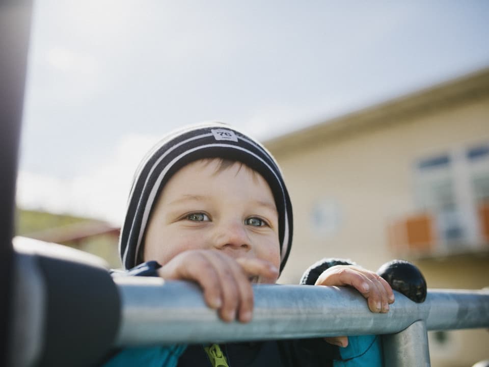 En pojke utomhus tittar fram bakom ett stålrör. Solen skiner och himlen är blå.
